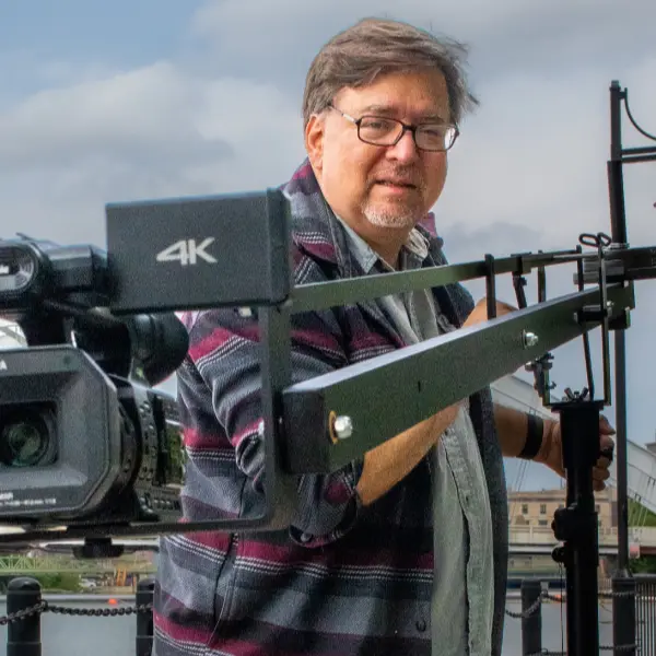 Paul Tracy operates a digital video camera, filming on location in Rochester, NY with the Genesee River in the background.