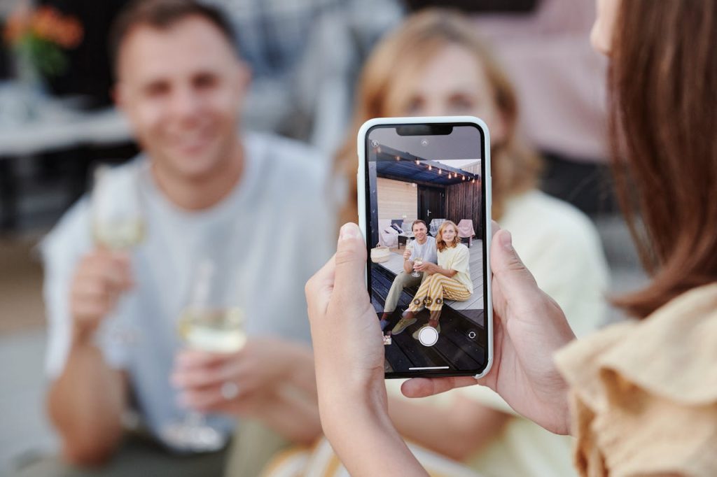 A person is taking a smartphone picture of a couple holding glasses of wine.