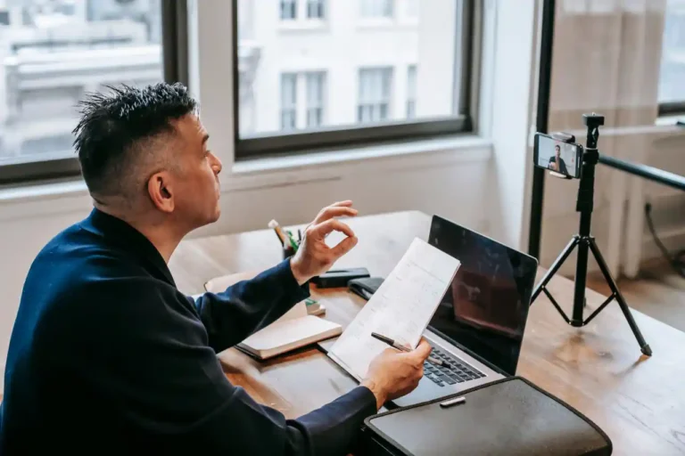 A man using his laptop and smartphone on a stand to make a video in an office setting. He's seated and holds his notes, making an emphasis gesture with his hand while speaking.