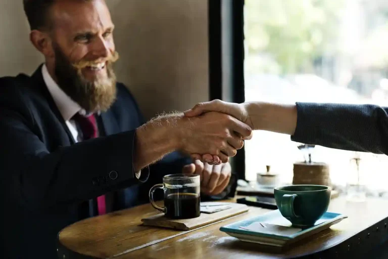 A smiling man wearing a suit and tie shakes the hand of someone off camera to the left. They're seated at a small cafe table, each with coffee mugs in front of them.