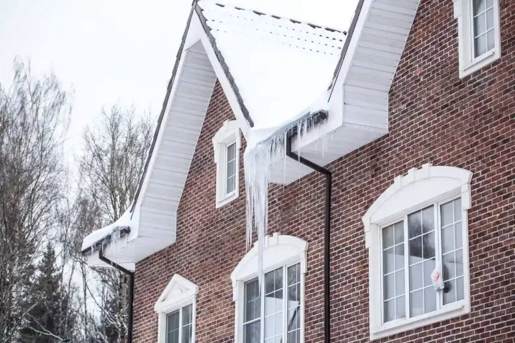 A brick house with snow-covered roof and ice damming above the gutter and large icicles hanging down.